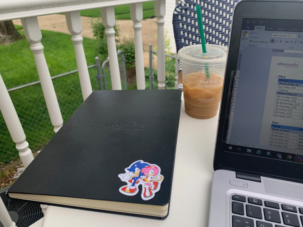 black bound notebook on a white table with Starbucks cup and a laptop. notebook has cartoon sticker on it.