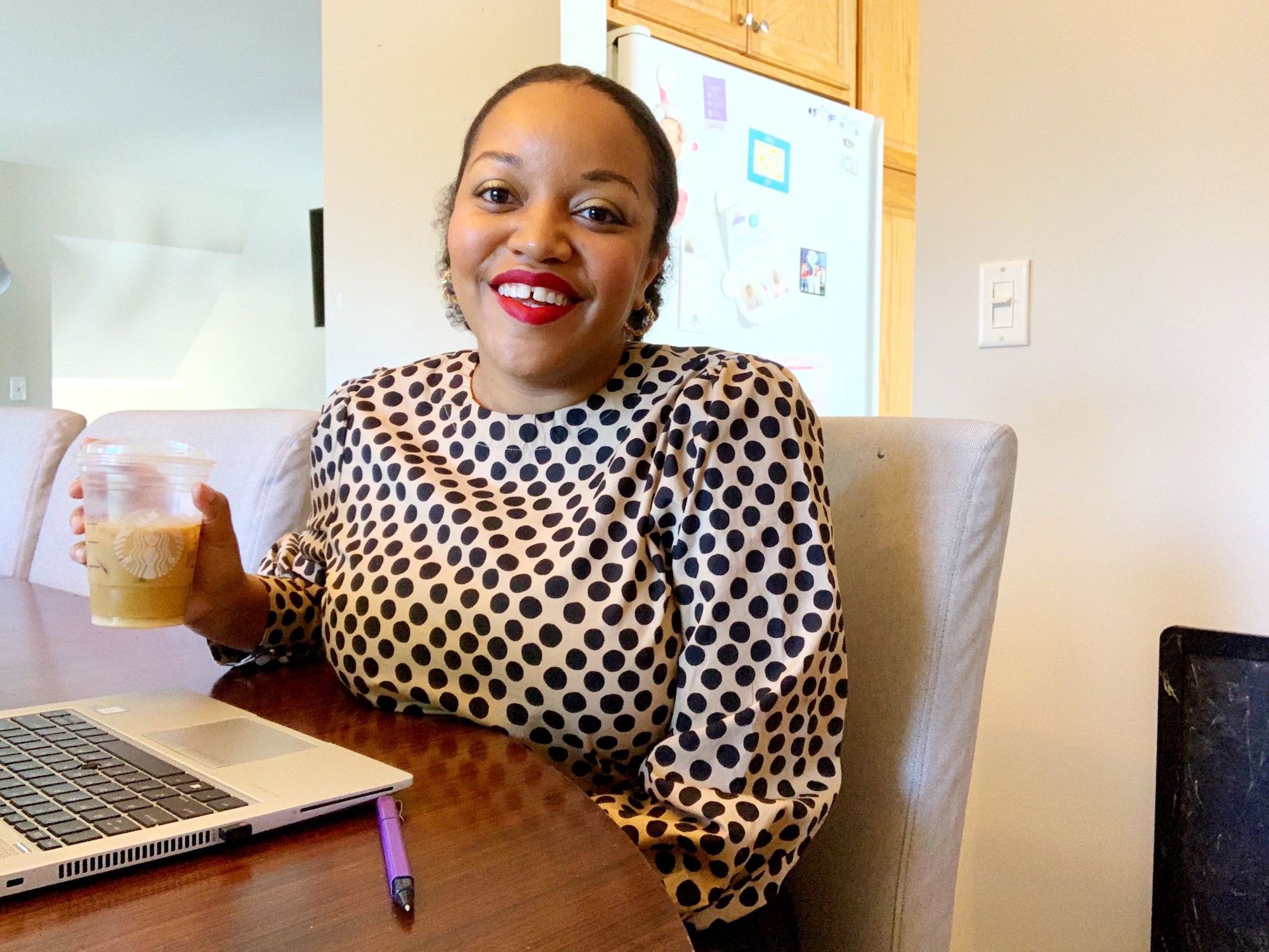 KBL smiling wearing a polka dot top and holding a cup of coffee. Sitting at the table with a laptop.