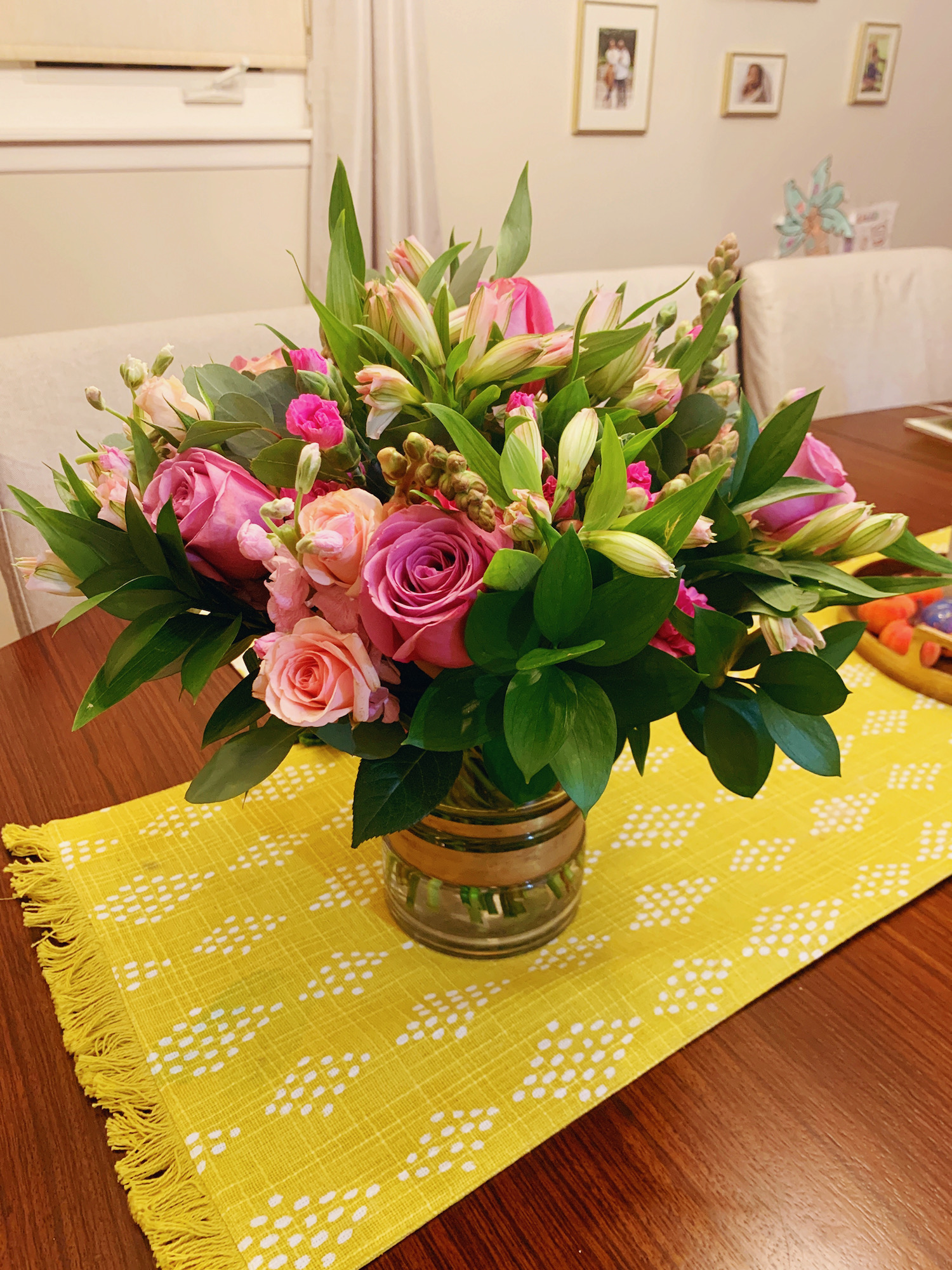 Large bouquet of flowers in different shade of pink inside of a gold vase. Vase on top of a yellow table linen.