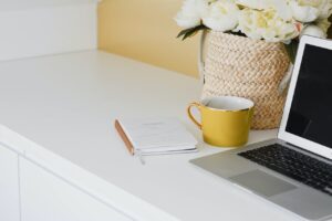 laptop on white counter with yellow coffee cup and notepad