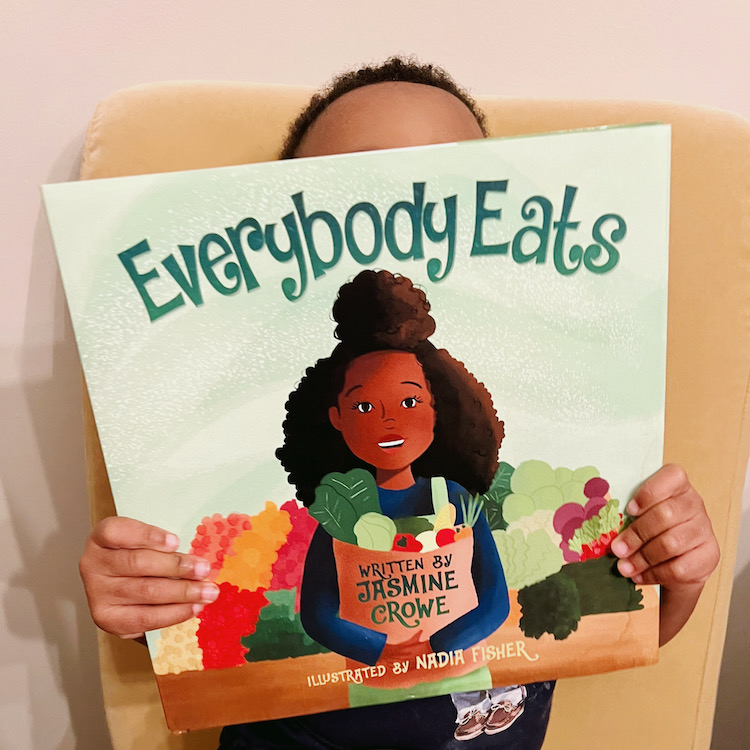 Black boy holding up the front of the book "Everyone Eats" by Jasmine Crowe. A children's book about food insecurity and hunger.