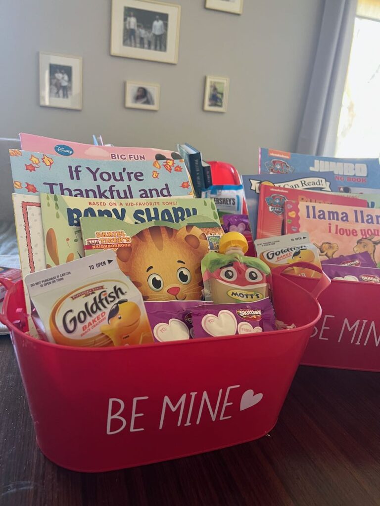 Two red gift bins with "Be Mine" on the front filled with Valentine's Day books for kids, candy, and goldfish crackers